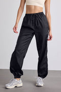 A model wearing black lightweight drawstring pants with elastic cuffs, paired with a beige crop top and silver sneakers, shown from the waist down against a light gray studio background.