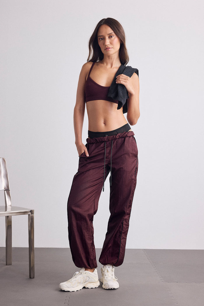 A woman modeling a matching burgundy activewear set consisting of a sports bra and jogger-style pants with a black waistband, holding a black towel over her shoulder, standing in a minimalist studio setting.